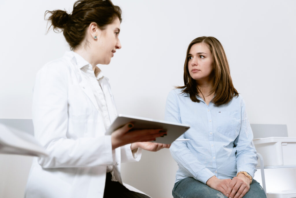 Clinician in a white coat holding a tablet speaking with a seated young woman in a light blue shirt.