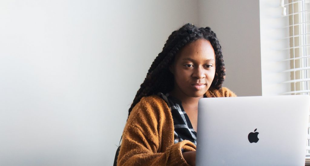 Person with long braided hair wearing a brown cardigan working on a silver MacBook laptop by a window