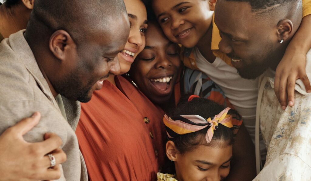 Family huddled closely, smiling and laughing while adults and children embrace; a girl with a colorful headband.