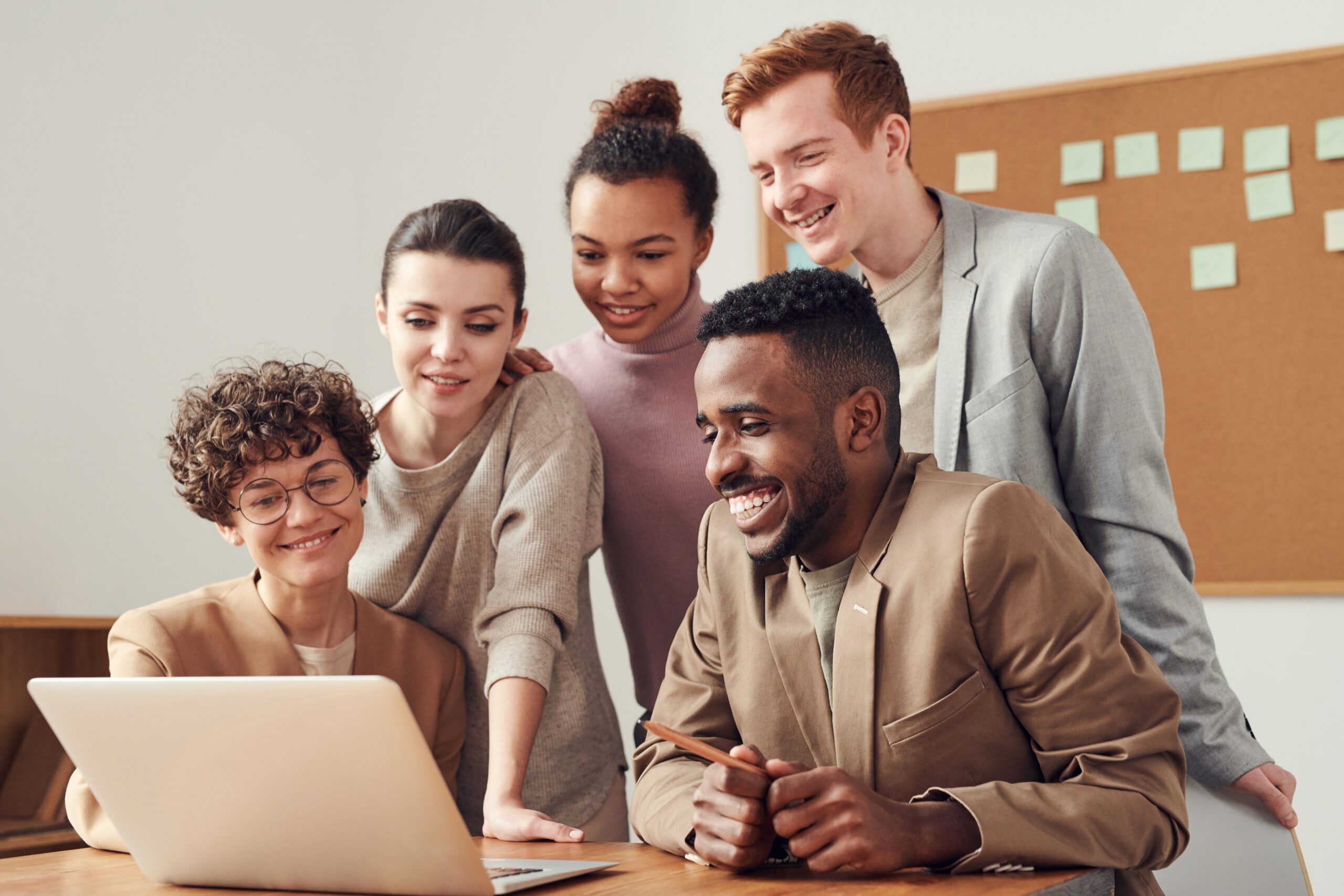 Five coworkers gathered around a laptop, smiling and reviewing work together with a corkboard of sticky notes behind them.