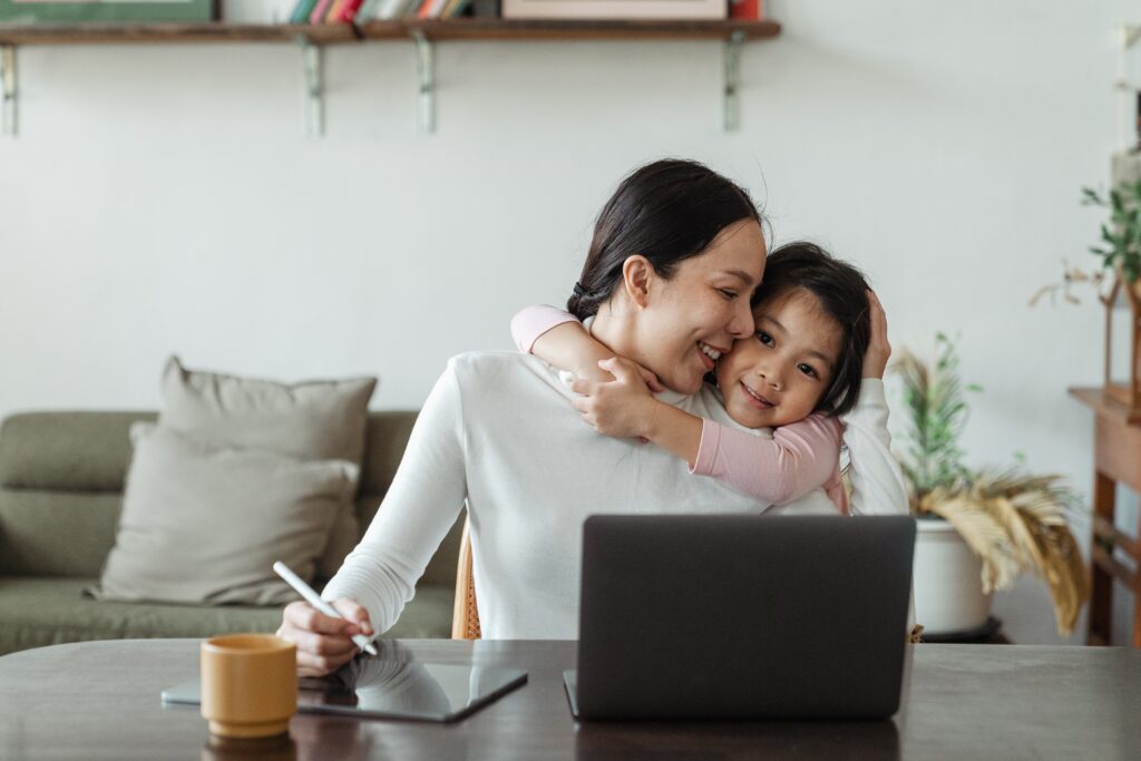Mother smiling as her young daughter hugs her from behind while she works on a laptop and tablet at home