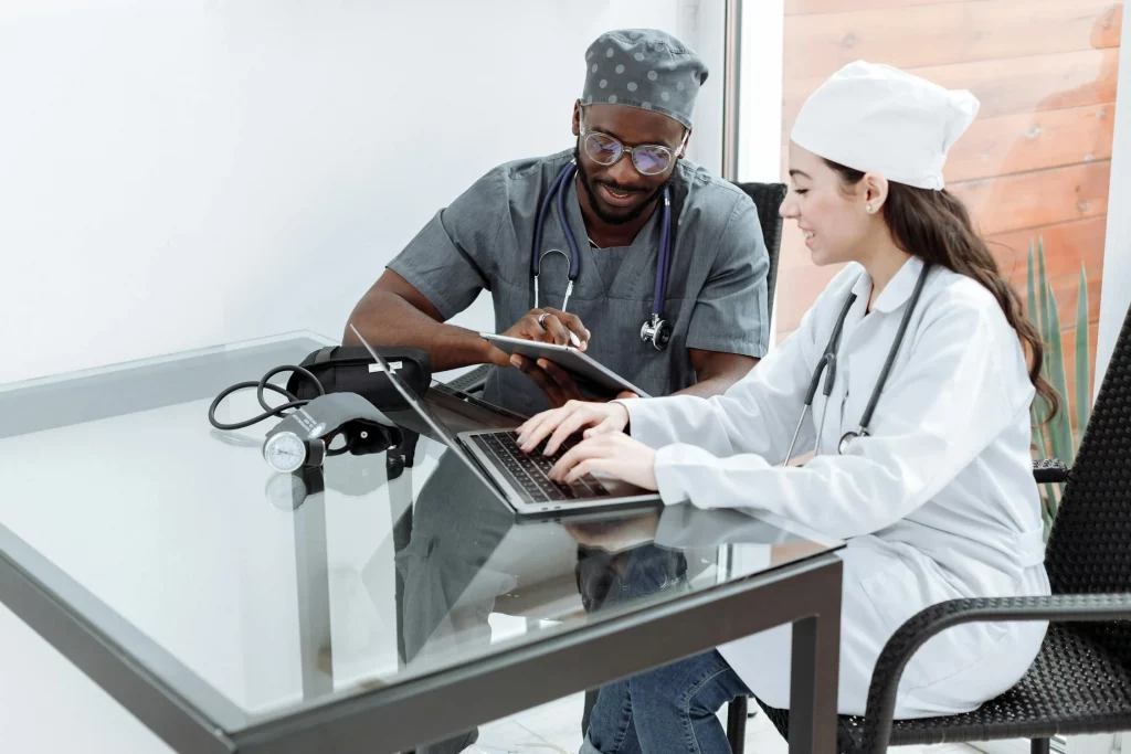 Two healthcare professionals in scrubs and lab coats work together at a glass desk, using a laptop and tablet, with a blood pressure monitor nearby.