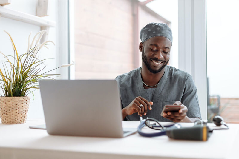 Smiling person in medical scrubs and cap at a desk using a smartphone, with a laptop and stethoscope visible