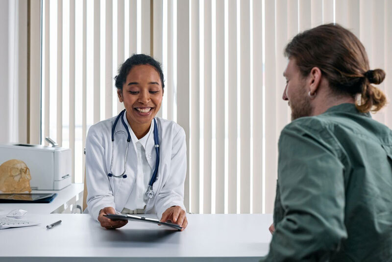 Smiling female doctor with stethoscope hands a tablet to a male patient across a clinic desk.